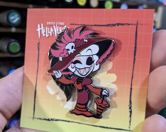 May include: A red and black enamel pin featuring a cartoon character with a red dress, black hair, and a large pink hat. The pin says "Into the Hellaverse" on the background card.