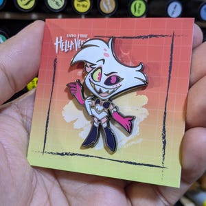 Puede incluir: Un pin de esmalte blanco y rosa con un personaje de dibujos animados con pelaje blanco, ojos amarillos y boca rosa. El personaje lleva un atuendo blanco y negro y sostiene un objeto rosa. El pin está en una tarjeta con el texto "Into the Hellaverse" en blanco y negro.
