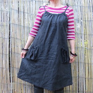 May include: A black linen dress with two pockets and a scoop neck. The dress has thin straps and is worn over a red and white striped shirt.