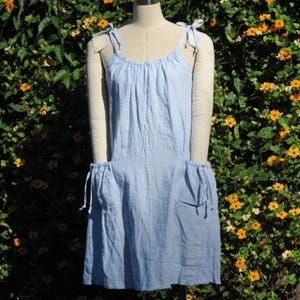May include: A light blue linen dress with tie straps and pockets. The dress is displayed on a mannequin in front of a backdrop of green foliage and yellow flowers. The dress has a relaxed, casual style.