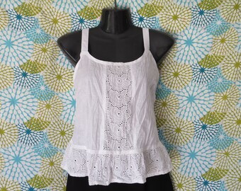 May include: White eyelet tank top with spaghetti straps and a ruffled hem. The top features a detailed eyelet design down the center and around the bottom. The top is displayed on a black mannequin against a patterned background.