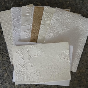 Puede incluir: Una colección de hojas de papel texturizadas con diseños en relieve. Los diseños incluyen ladrillos, flores y motivos de remolinos. Los papeles son de color blanco y marrón claro. Las hojas están dispuestas en forma de abanico.