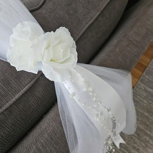 May include: White floral chair decoration with two large fabric flowers, tulle, and a satin ribbon adorned with small pearls. The decoration is draped over the arm of a gray upholstered chair.