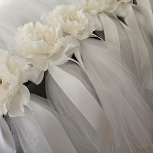 May include: A close-up of a white tulle skirt adorned with large, cream-colored peony flowers and satin ribbons. The skirt is layered, creating a soft, flowing effect. The ribbons hang down, adding to the delicate design.