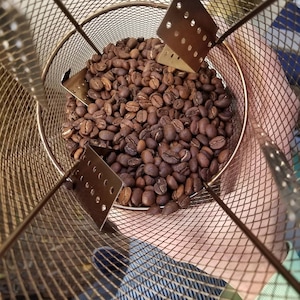 Puede incluir: Primer plano de una cesta de tostado de metal llena de granos de café tostados. Los granos son de color marrón oscuro y tienen una superficie brillante. La cesta está hecha de una malla fina y tiene un asa en el lateral.