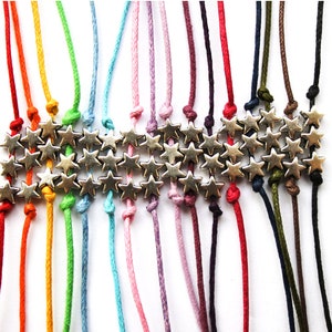 Può includere: Un set di 18 braccialetti colorati in corda con perline a forma di stella argentate. I braccialetti sono legati insieme in alto con un nodo.