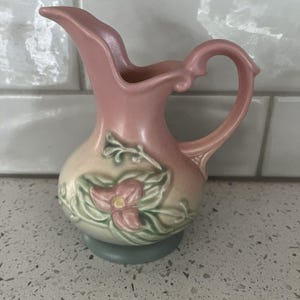 May include: A small, pink ceramic pitcher with a curved spout and handle. The pitcher features a floral design in pink, green, and cream colors. The base is a muted blue-green color. The pitcher is likely vintage or antique.