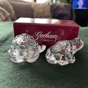 May include: Two clear crystal figurines: a turtle and a frog. The turtle has a textured shell, and the frog is in a sitting position. A red box with the Gorham logo is in the background.