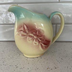 May include: A vintage ceramic creamer with a floral design. The creamer has a rounded body with a handle and a spout. The top is a light blue, transitioning to a creamy yellow body with a raised red floral design. The handle is yellow.