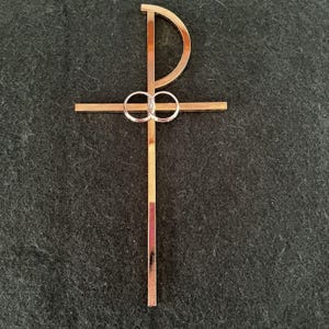May include: A metallic rose gold Chi Rho symbol featuring a vertical bar intersected by a horizontal bar and a curved top section. Two interlocking circular rings are positioned at the center where the horizontal and vertical bars meet.