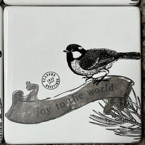 May include: Black and white ceramic tile coaster with a bird perched on a branch. The branch has a banner that reads "joy to the world". A vintage stamp with the text "Seasons Greetings 1915" is in the upper left corner.