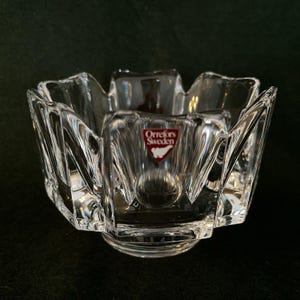 May include: Clear crystal bowl with a unique, multi-faceted design. The bowl has a flared, petal-like rim and a round base. A red and white label with the text "Orrefors Sweden" is visible on the front.