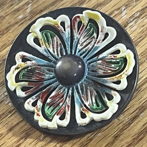 May include: A circular button featuring a dark gray base and a raised, white, flower-shaped overlay. The petals are accented with streaks of green, red, and yellow paint, and a smooth, dark gray circular dome sits at the center of the flower.