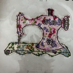 May include: A fabric sewing machine applique patch featuring a pink floral pattern on the main body and a cream-colored floral pattern on the base. The design is outlined with black thread embroidery, highlighting the machine's shape and details.
