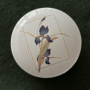 May include: A round, white ceramic dish with a floral design. The design features a blue iris flower with gold accents and a gold geometric frame. The background has vertical gray stripes.