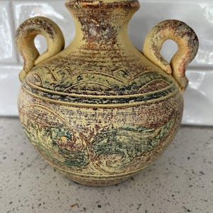 May include: A small, yellow ceramic vase with two handles and a brown, green, and yellow painted design.