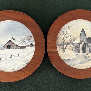 May include: Two circular wall plaques featuring winter landscape paintings in wooden frames. The left plaque shows a snow-covered barn and fence. The right plaque depicts a stone water mill in a snowy setting. Both paintings are signed D. Cuddy.