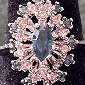 May include: A close-up of a silver ring with a large, dark blue, marquise-cut gemstone at the center. The ring is surrounded by smaller, pink and gray gemstones, creating a floral or snowflake design. The ring is set against a dark background.