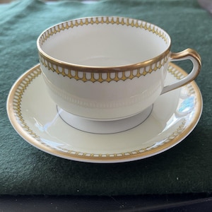 May include: A white teacup and saucer set with gold trim. The cup has a curved handle and a scalloped gold border. The saucer also has a gold border. The set is on a green surface.