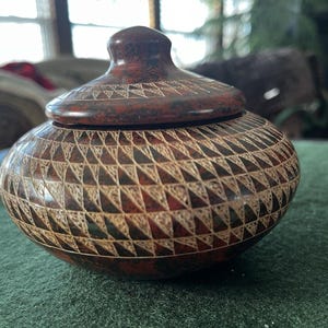 May include: A decorative, lidded jar with a dark reddish-brown finish. The jar's surface is intricately carved with a repeating geometric pattern of triangles. The lid has a rounded knob and a similar carved design. The jar sits on a green surface.