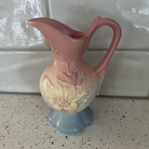 May include: A small, vintage-style pitcher in shades of pink, yellow, and blue. The pitcher features a floral design on the front and a curved handle. The base is a light blue color. This decorative piece is likely ceramic.
