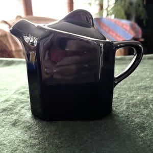 May include: A glossy black ceramic creamer or small pitcher featuring a geometric, angular body with a distinct spout, a curved handle on the side, and a matching lid with a raised, pointed top section, reflecting light on its smooth surface.