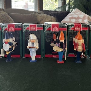 May include: A collection of Hallmark Keepsake Christmas ornaments featuring various wooden figures. The ornaments include a soldier, a musician, a chef, an artist, a Santa-like figure, and a bearded man. Each ornament is in its original box.