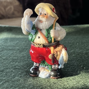 May include: A figurine of a jolly fisherman, wearing a yellow hat, green vest, and red pants. He holds a fishing rod and two fish. The figurine has a detailed design with a white beard and black boots.