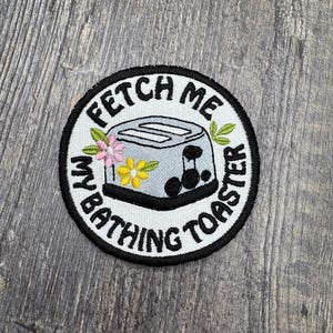 May include: Embroidered patch with a black border and the text "FETCH ME MY BATHING TOASTER". The patch features a gray toaster with pink and yellow flowers and green leaves.