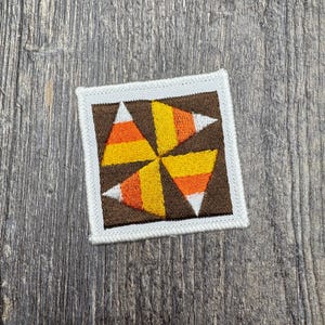 May include: A square embroidered patch featuring a pinwheel design composed of four candy corn shapes. Each candy corn segment displays horizontal stripes of white, orange, and yellow. The design is set against a dark brown background with a white border.