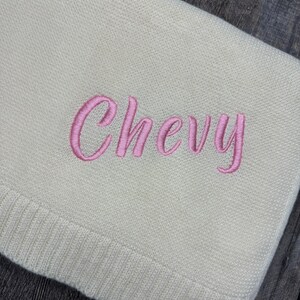 K&ouml;nnte beinhalten: Eine cremefarbene Strickdecke mit dem in kursiver rosa Schrift eingestickten Namen Chevy. Der Stoff hat eine feine, gleichm&auml;&szlig;ige Textur mit einem gerippten Rand an der Unterseite, was ein schlichtes Design f&uuml;r diesen Textilartikel ergibt.