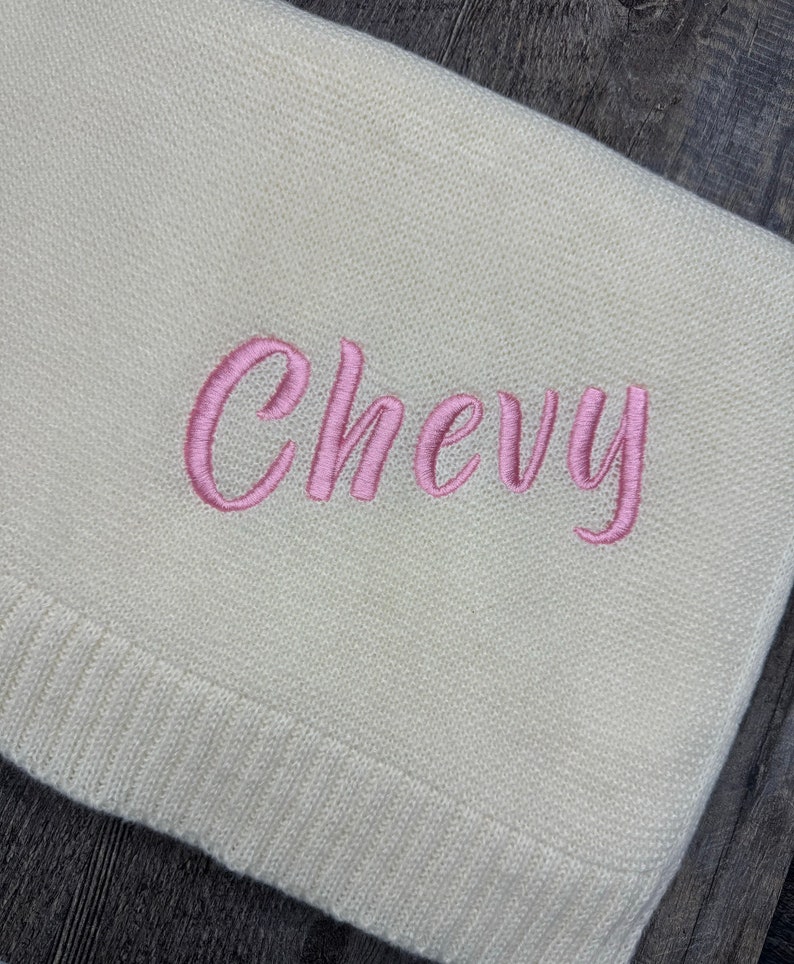 K&ouml;nnte beinhalten: Eine cremefarbene Strickdecke mit dem in kursiver rosa Schrift eingestickten Namen Chevy. Der Stoff hat eine feine, gleichm&auml;&szlig;ige Textur mit einem gerippten Rand an der Unterseite, was ein schlichtes Design f&uuml;r diesen Textilartikel ergibt.