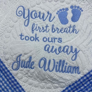 May include: A white baby quilt with blue gingham trim. The quilt is embroidered with the text "Your first breath took ours away Jude William" and two blue footprints.