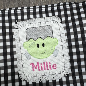 May include: A white fabric patch with a green Frankenstein face and the name Millie embroidered in pink. The patch has a frayed edge and is on a black and white checkered background.