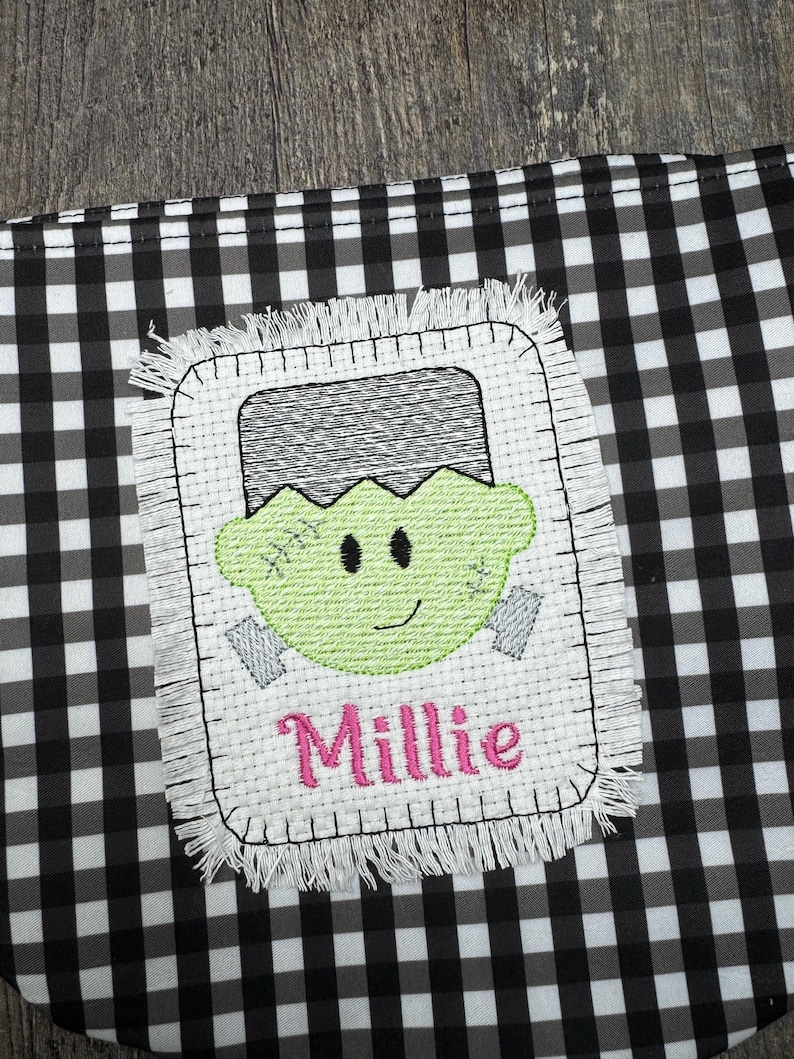 May include: A white fabric patch with a green Frankenstein face and the name Millie embroidered in pink. The patch has a frayed edge and is on a black and white checkered background.