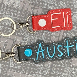 May include: Two personalized leather keychains with metal rings and clasps. The top keychain is red with white stitching and the name "Eli" embroidered in white thread. The bottom keychain is gray with blue stitching and the name "Austin" embroidered in blue thread.