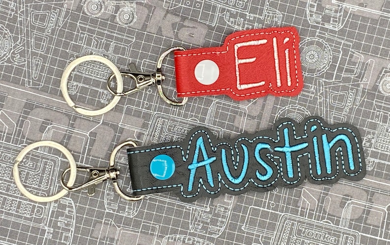 May include: Two personalized leather keychains with metal rings and clasps. The top keychain is red with white stitching and the name "Eli" embroidered in white thread. The bottom keychain is gray with blue stitching and the name "Austin" embroidered in blue thread.