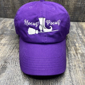 May include: Purple baseball cap with white embroidery of a witch's boot, broom, and the text "Hocus Pocus".