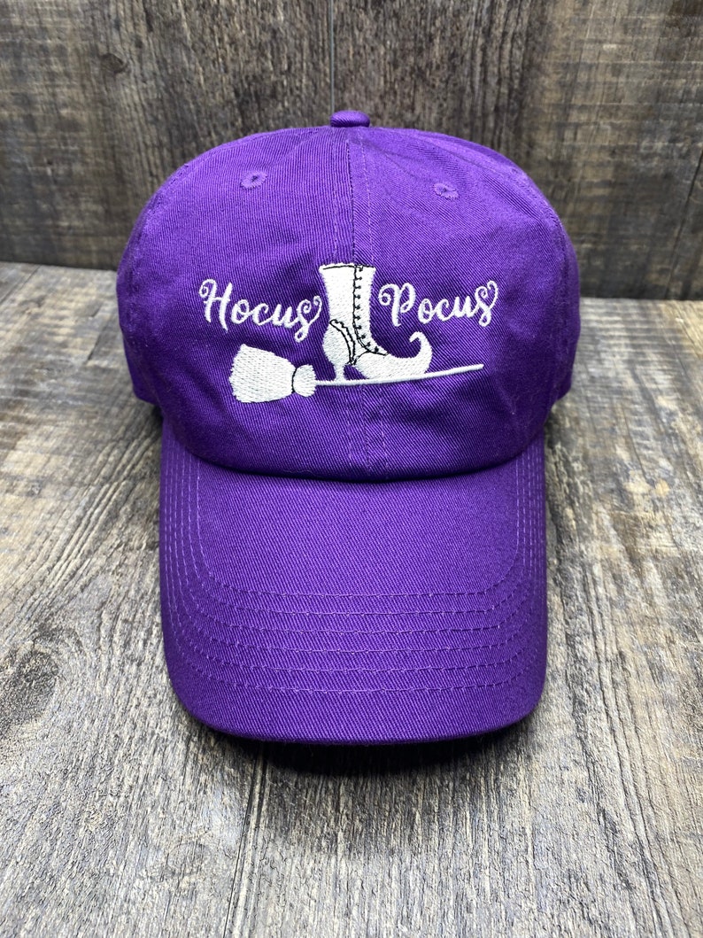 May include: Purple baseball cap with white embroidery of a witch's boot, broom, and the text "Hocus Pocus".