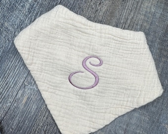 May include: Cream-colored baby bib with a purple embroidered letter "S". The bib is made of a textured fabric and is laying on a wooden surface. The bib is a bandana style, designed to catch drool and spills.