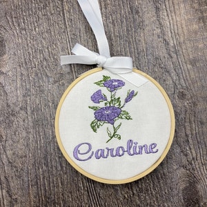 May include: A white fabric hoop with a wooden frame, featuring an embroidered purple flower design and the name "Caroline" in cursive script. The hoop is attached to a white ribbon.