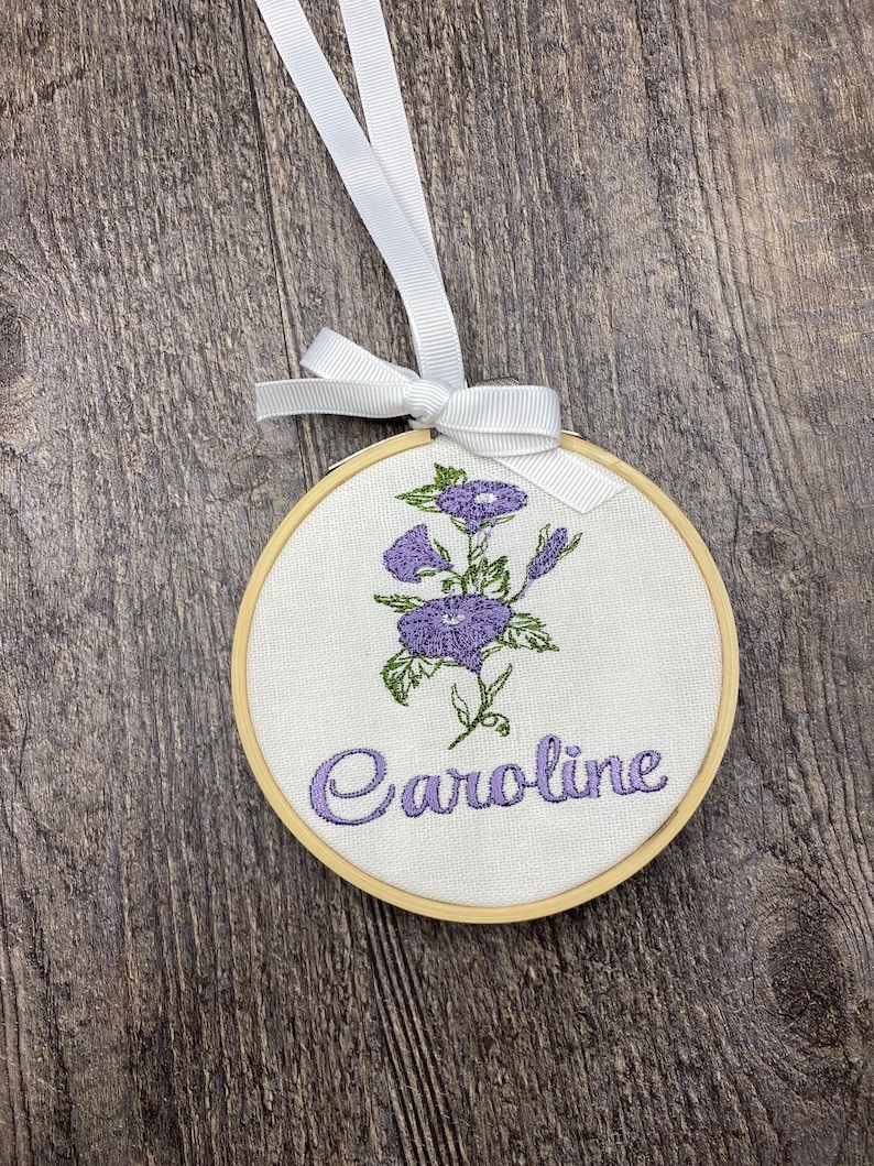 May include: A white fabric hoop with a wooden frame, featuring an embroidered purple flower design and the name "Caroline" in cursive script. The hoop is attached to a white ribbon.