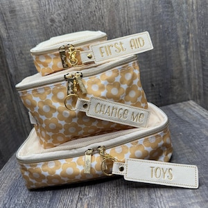 May include: A stack of three zippered cosmetic bags with a daisy pattern in yellow and white. Each bag has a cream-colored trim and a gold zipper. The top bag has a tag that says "FIRST AID". The middle bag has a tag that says "CHANGE ME". The bottom bag has a tag that says "TOYS".