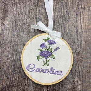 May include: A white embroidery hoop with a purple flower design and the name "Caroline" embroidered in purple thread.