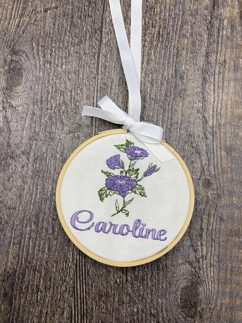 May include: A white embroidery hoop with a purple flower design and the name "Caroline" embroidered in purple thread.