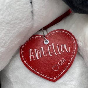 May include: A red heart-shaped tag with the name "Amelia" in white script. The tag is attached to a plush toy. The tag also has the initials "GM" and a small heart embroidered on it.