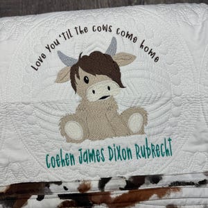 May include: White quilted baby blanket with a brown cow applique and the text "Love You 'Til the Cows Come Home" and the name "Cohen James Dixon Rubrecht" embroidered in teal.