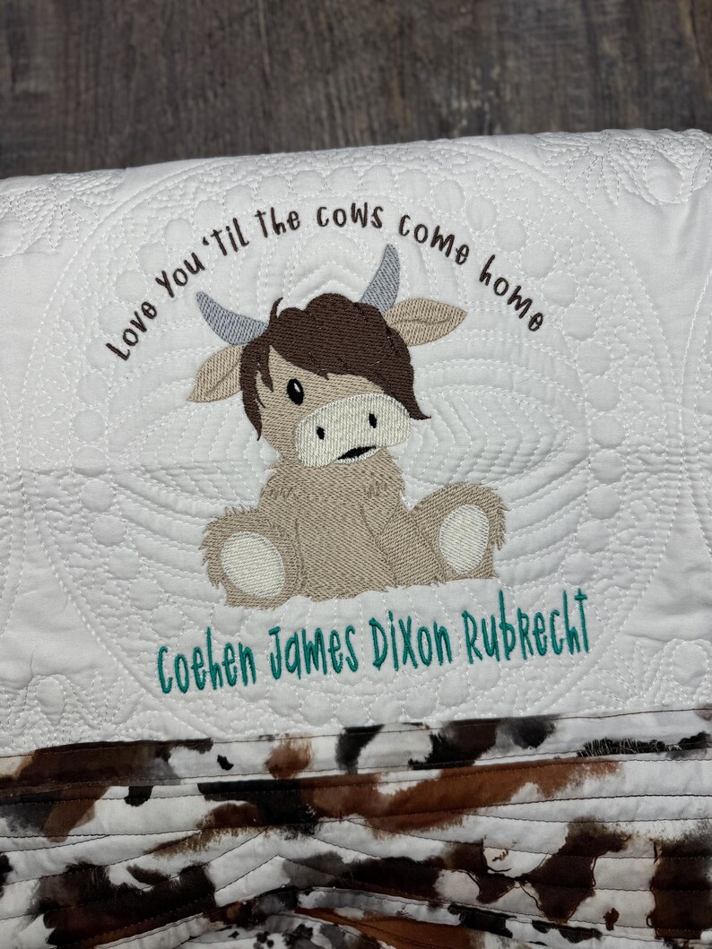 May include: White quilted baby blanket with a brown cow applique and the text "Love You 'Til the Cows Come Home" and the name "Cohen James Dixon Rubrecht" embroidered in teal.