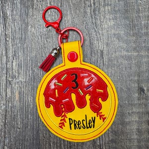 May include: A yellow and red donut-shaped keychain with the number "3" and the name "Presley" embroidered on it. The keychain has a red tassel and a red clasp.