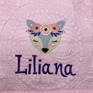 K&ouml;nnte beinhalten: Eine rosa gesteppte Babydecke mit einem gestickten Hirsch mit Blumen und dem Namen "Liliana" in lila Stickerei.